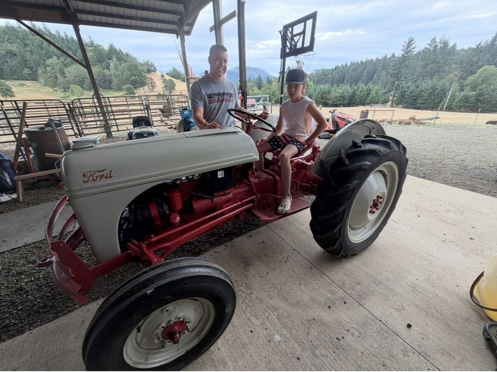 A man and a boy sitting on a restored gray-and-red vintage Ford tractor under a pole barn, with green hills behind