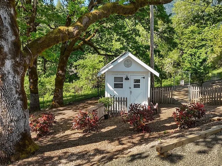 Small white chicken coop with a picket fence under large oak trees